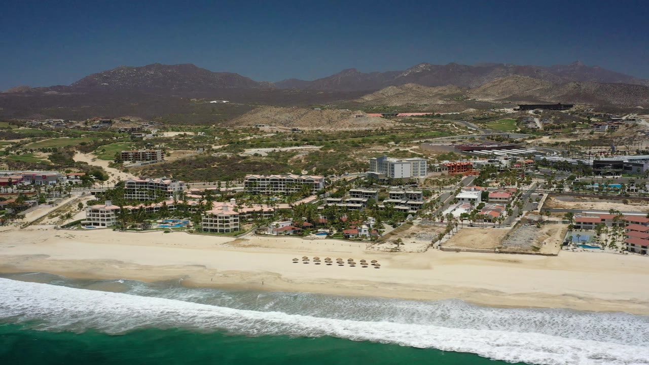 Aerial view in San Jose del Cabo with a white wave band crossing the lower frame, umbrellas on the sand, clustered resort buildings inland, and no visible people.