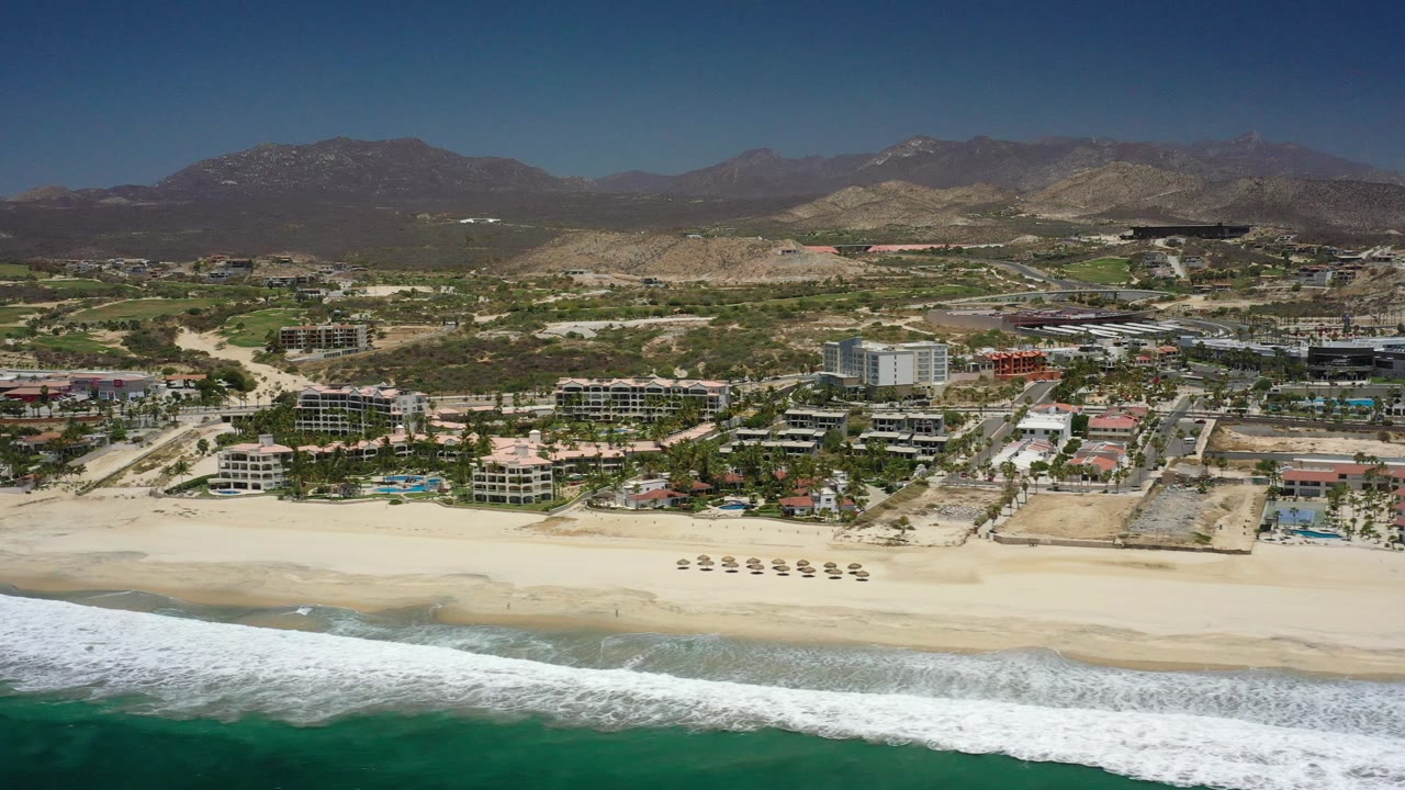 High coastal aerial with beach, surf, hotels, roads, and dry mountains.