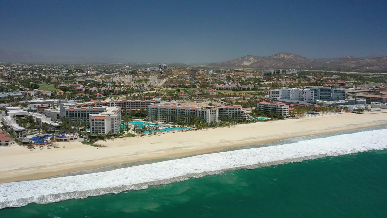 Wide opening frame over San Jose Del Cabo with green-blue ocean in the foreground, white surf along a sandy beach, resort buildings and pools behind the shore, inland city blocks, distant hills, and no visible people.