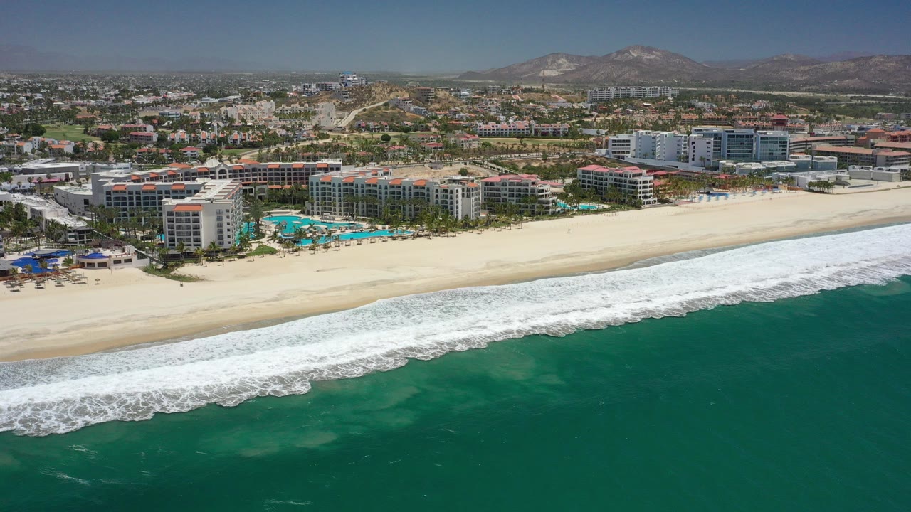 Aerial view of dark green ocean, white surf, sandy beach, resort buildings, and distant hills.