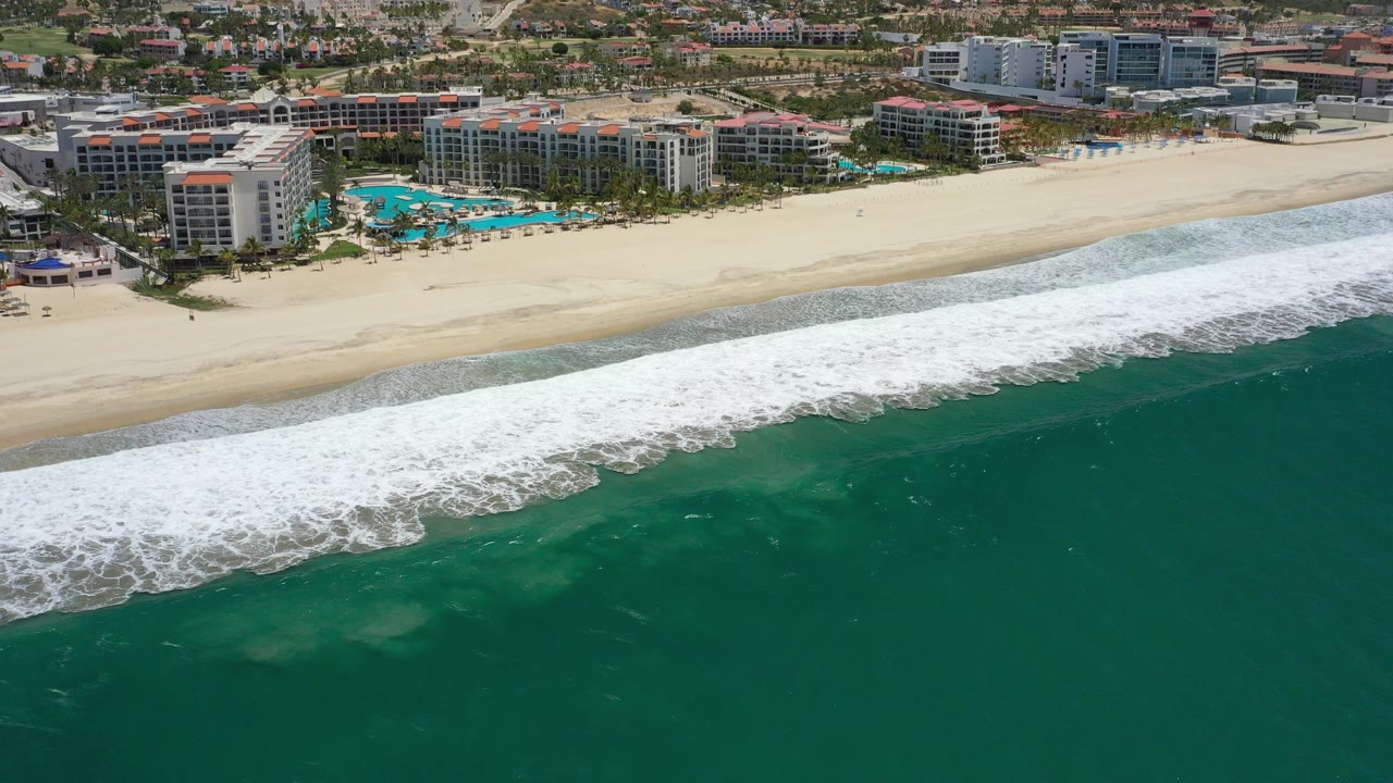 Aerial view off San Jose del Cabo, Mexico with turquoise water in the foreground, white surf crossing the beach, resort buildings beyond the sand, and no visible people.