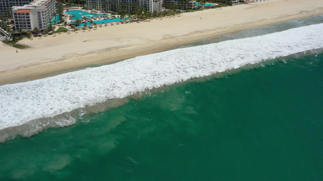 Aerial view of white surf breaking onto a wide tan beach beside green ocean.