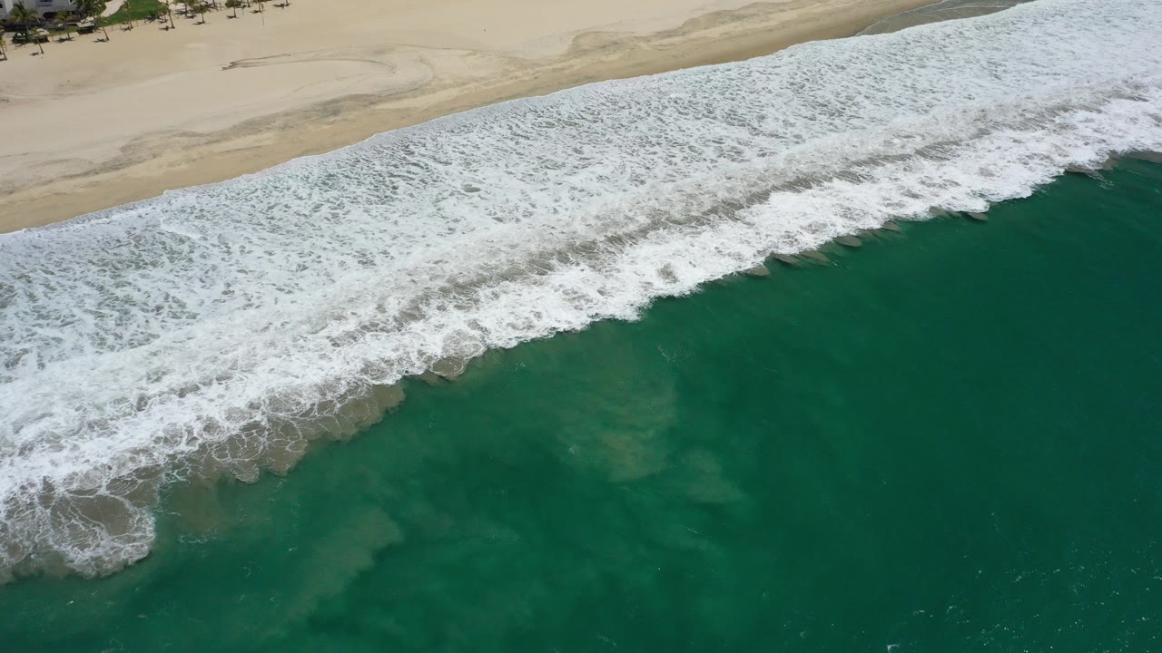 Overhead view of dark green ocean water and white surf approaching a sandy beach in San Jose del Cabo, Mexico, with no visible people.