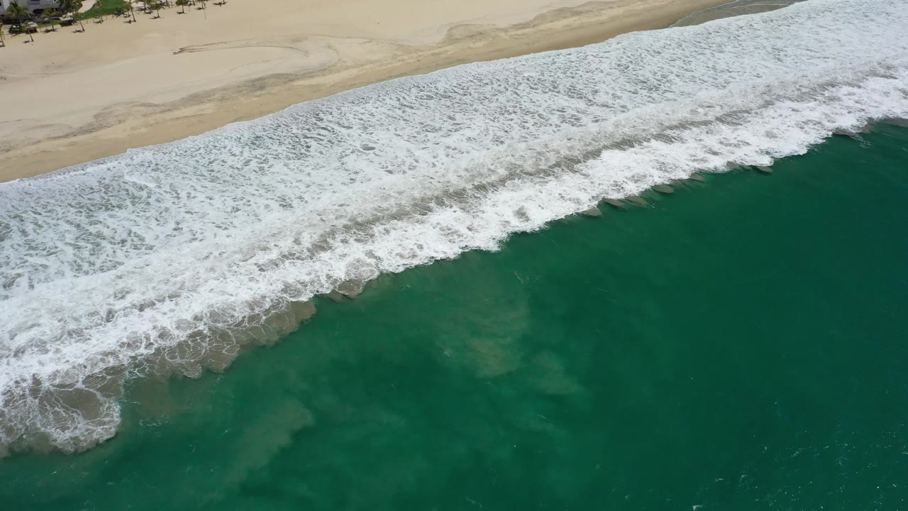 Aerial frame of white foam spreading over dark green water below a pale sandy shoreline in San Jose del Cabo, Mexico, with no visible people.