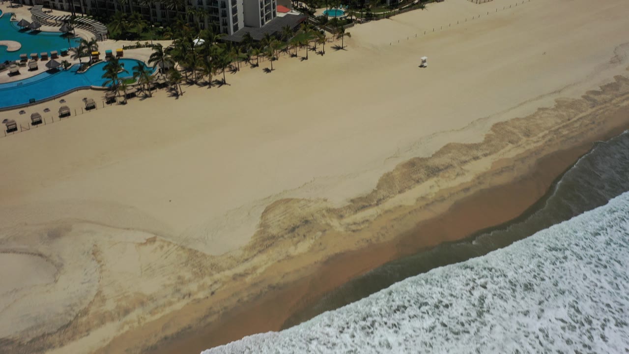 A high aerial frame over a San Jose del Cabo resort beachfront shows turquoise pools at upper left, palm trees along the sand, a broad beach, and white surf at the lower right.