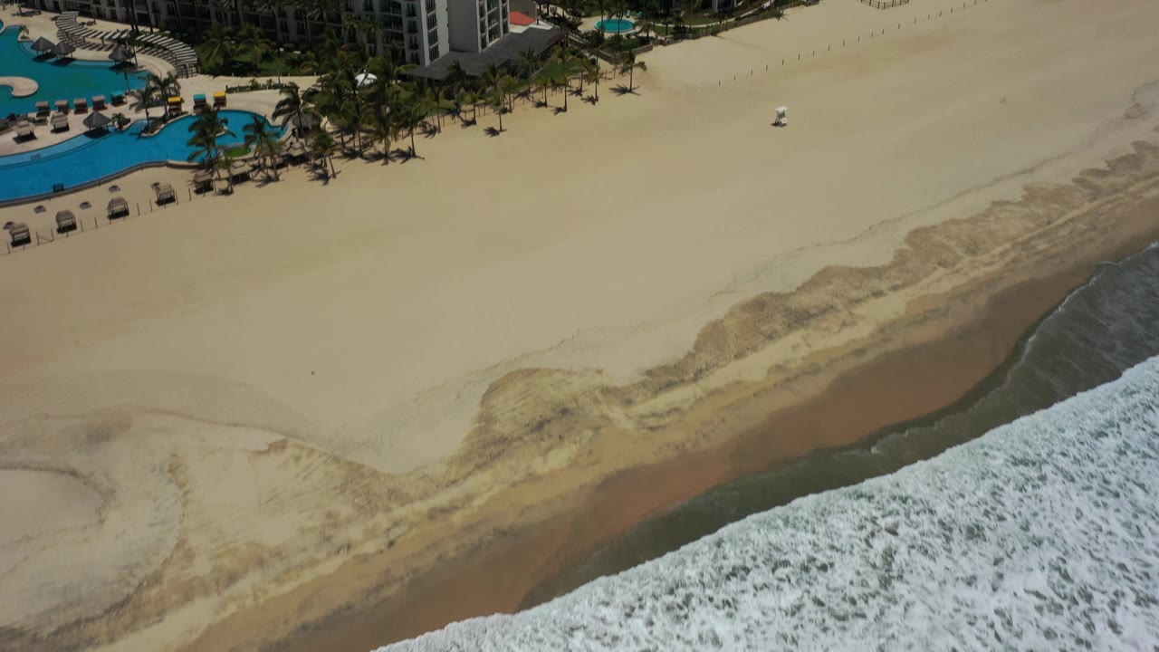 The San Jose del Cabo beach fills the center of the frame, with resort buildings and pool decks near the top and a band of foamy ocean water along the lower right.