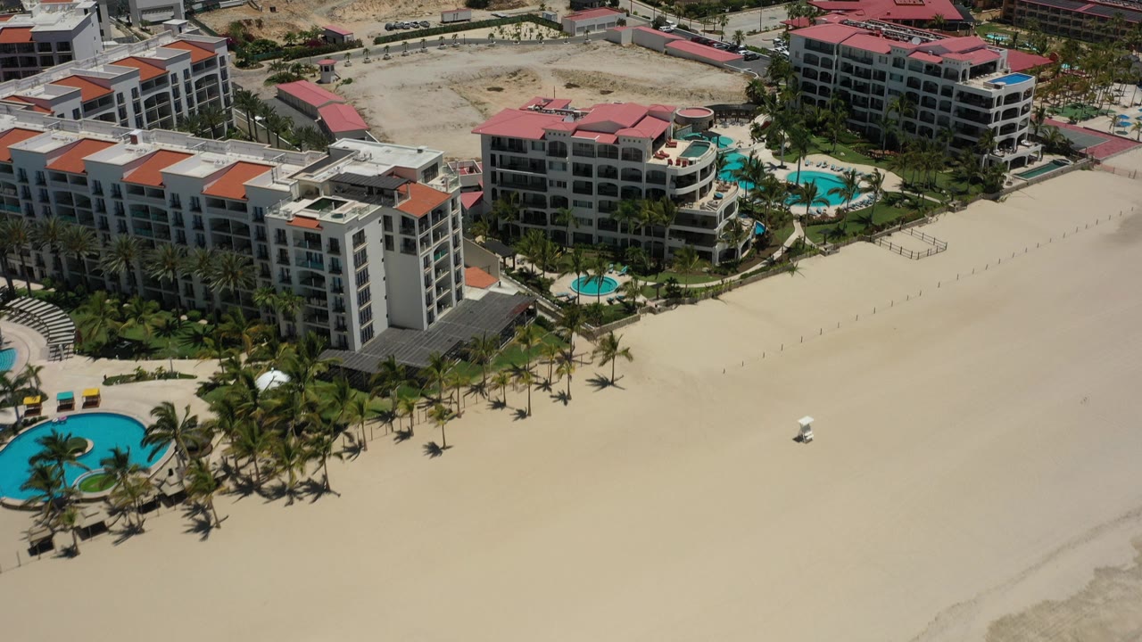 Opening aerial frame over San Jose del Cabo hotel buildings with red and white roofs, turquoise pools, palm landscaping, wide sandy beach, inland lots, and no visible people.