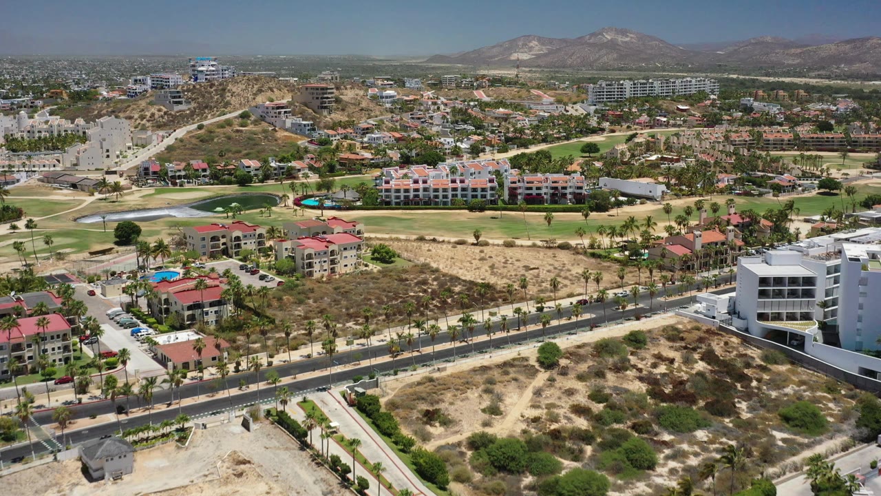 Aerial view in San Jose Del Cabo, Mexico, with a white modern building near the lower right, red-roofed complexes, green fairways, tan vacant land, distant hills, and no visible people.