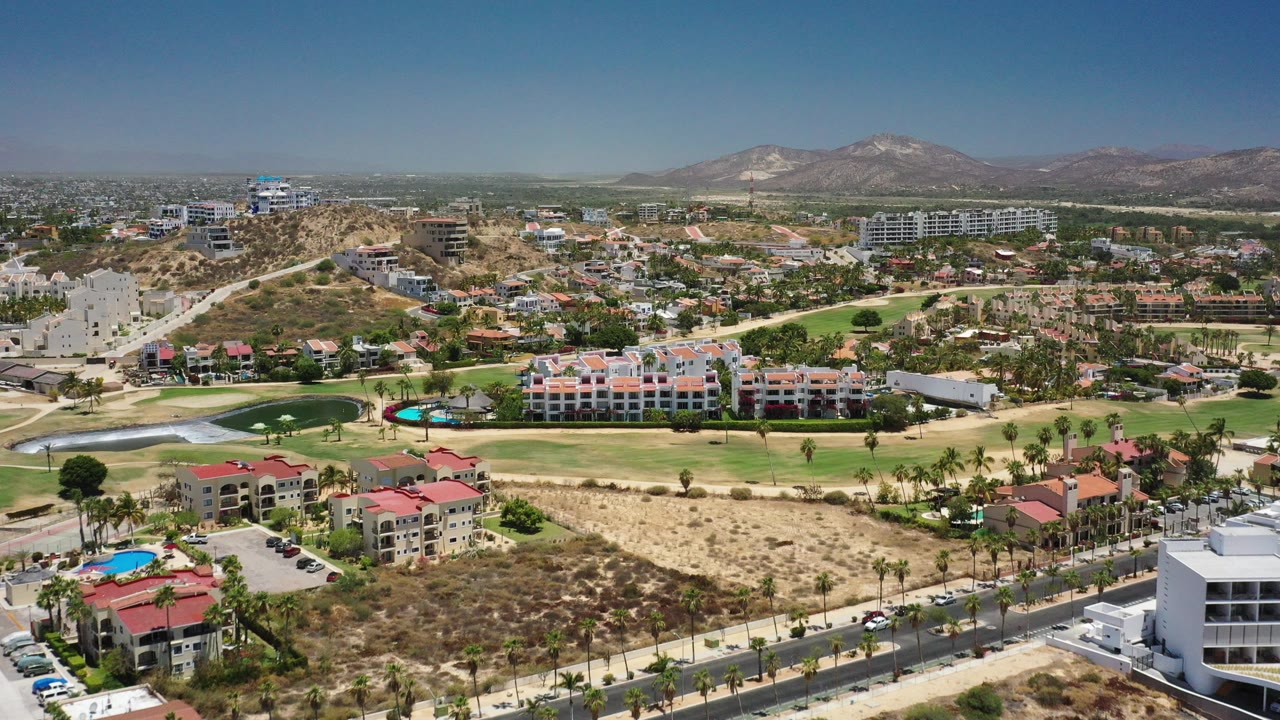Wide aerial view of housing, golf greens, dry lots, and mountains under blue sky.