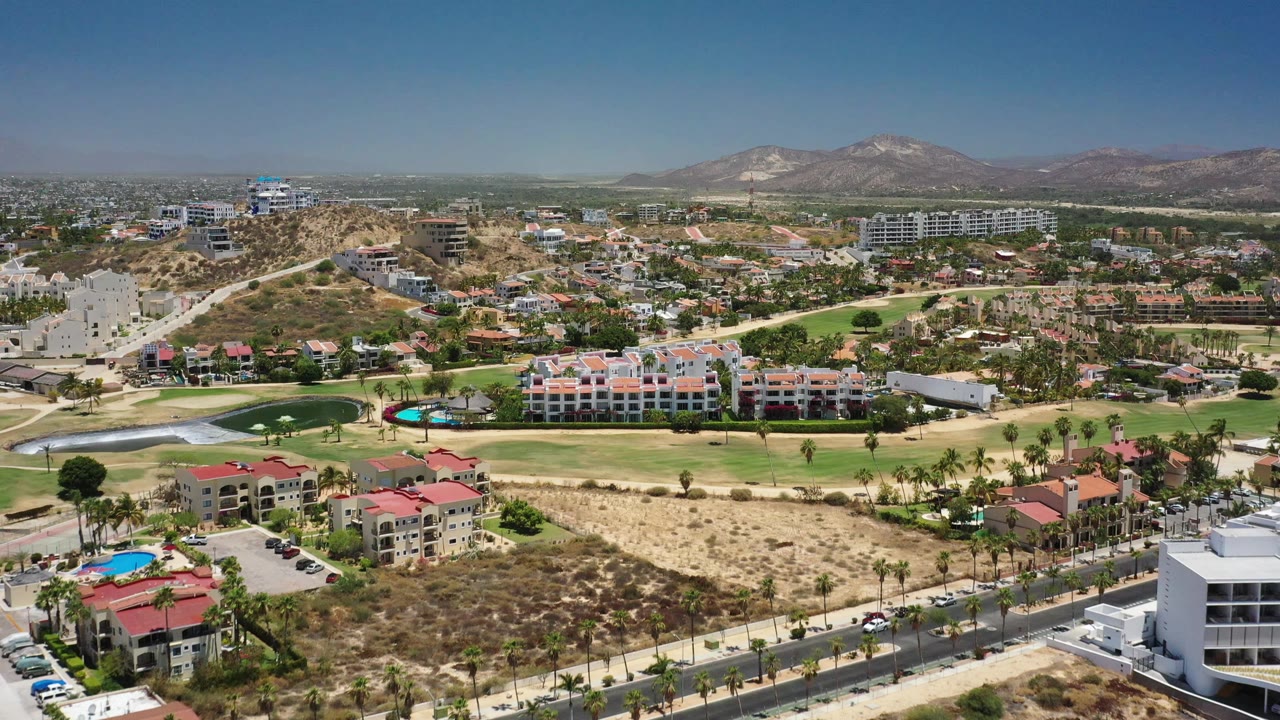 Wide aerial frame of San Jose del Cabo showing curving golf greens and sand-colored paths in front of apartment blocks, dry hillside development, mountains, and no visible people.
