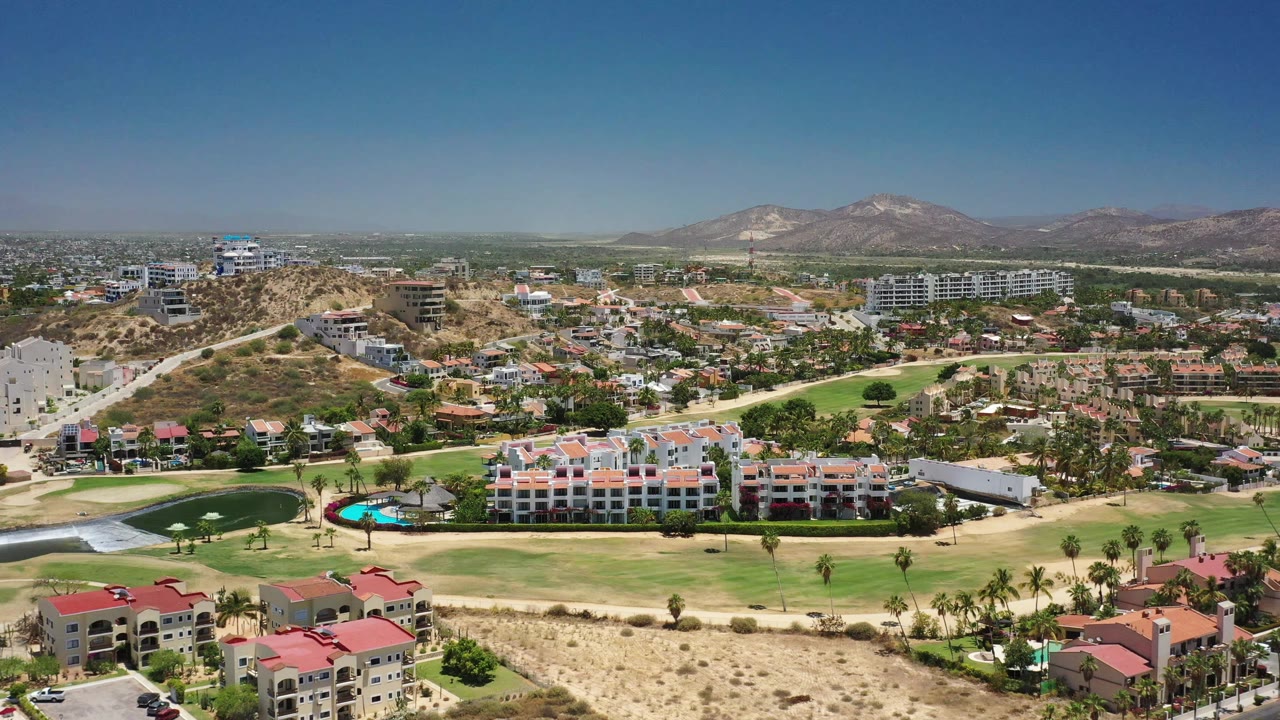 Second aerial frame in San Jose Del Cabo holds a wide view of low-rise homes, a curved golf green, a bright pool, roads, hillside construction, and mountain ridges, with no visible people.