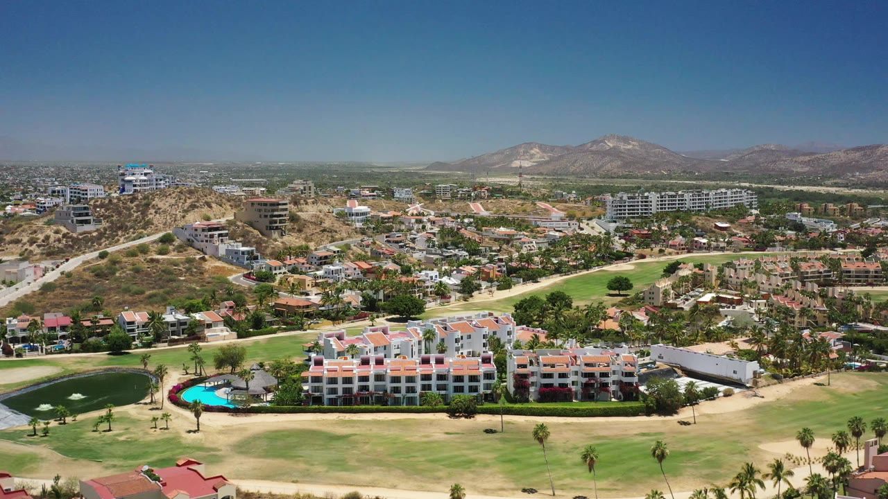 San Jose del Cabo aerial view one second in, with a turquoise pool near resort buildings, manicured fairways below, residential streets behind, dry hills, mountains, and no visible people.