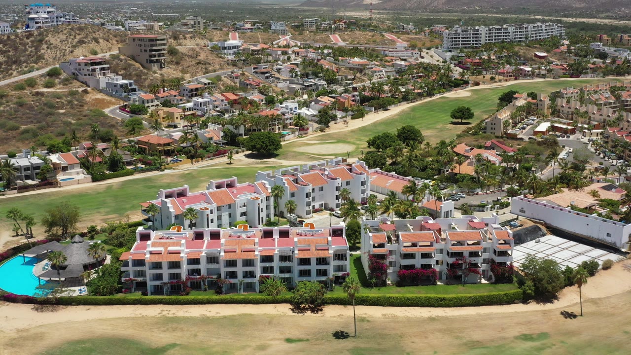 Opening aerial frame over San Jose Del Cabo showing white tourist buildings with red roofs, a bright blue pool at left, golf fairway, roads, hillside homes, and no visible people.