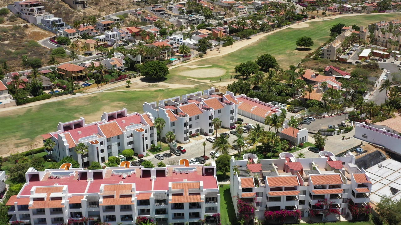 Aerial view of white buildings with red roofs beside green fairways and parked cars.
