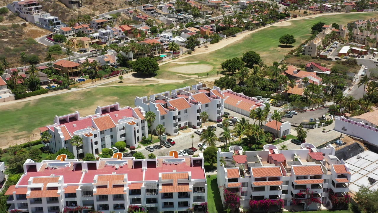 Aerial frame in San Jose del Cabo with the red-roofed building complex filling the lower frame, parked cars at right, fairways behind, and no visible people.