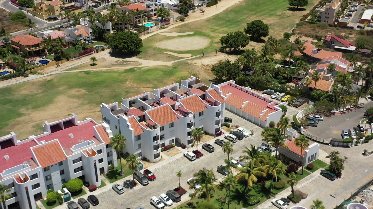 Overhead frame in San Jose del Cabo showing red-roofed white apartments, a parking area, palm trees, a road curve, and a golf fairway behind them, with no visible people.