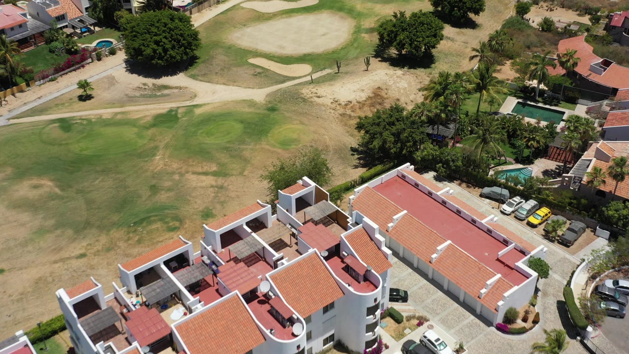 High aerial view in San Jose del Cabo, Mexico, showing white homes with terracotta roofs beside patchy green and tan landscaped ground, trees, pools, parked cars, and no visible people.