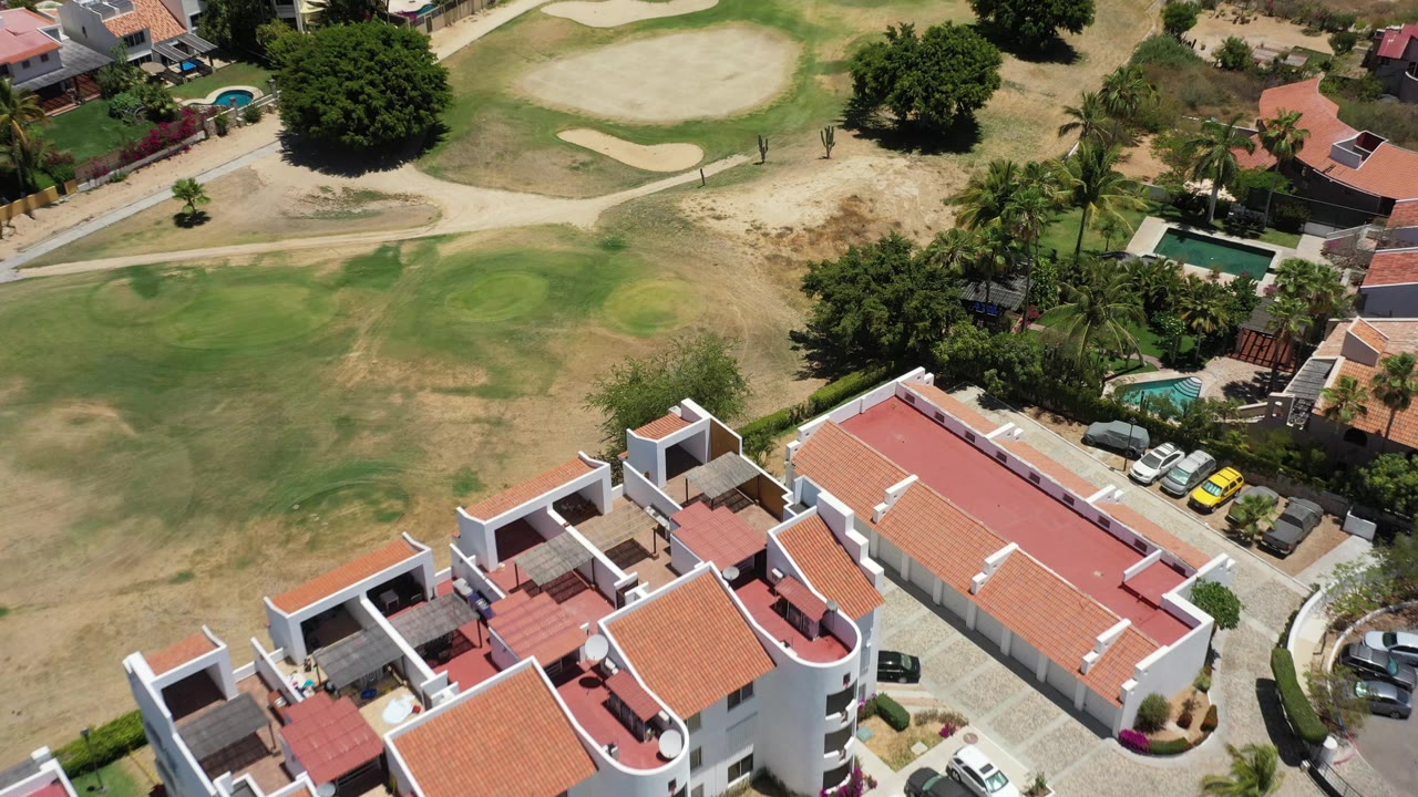A second aerial frame over San Jose del Cabo, Mexico, shows the residential roofs lower in frame, parked cars near a road, small blue-green pools, palms, and no visible people.
