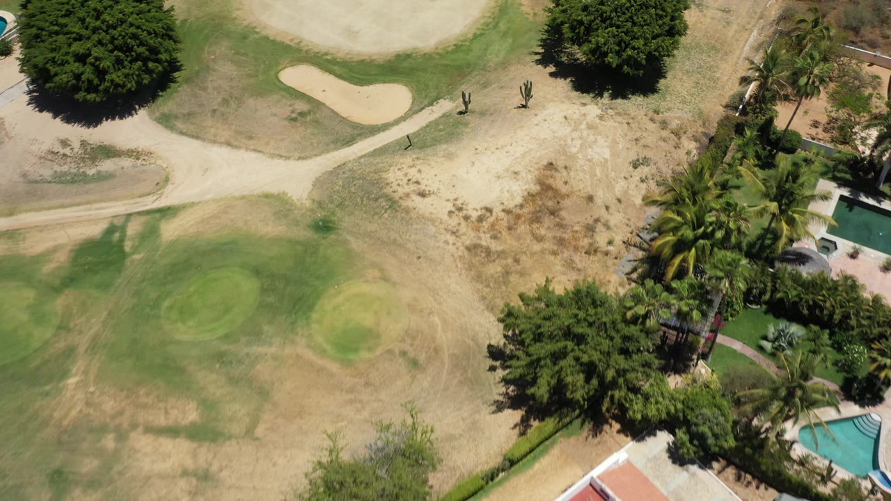 San Jose del Cabo aerial frame shows a dry golf-course area with two green patches, a sand trap near the top, trees casting shade, palms, and no visible people.