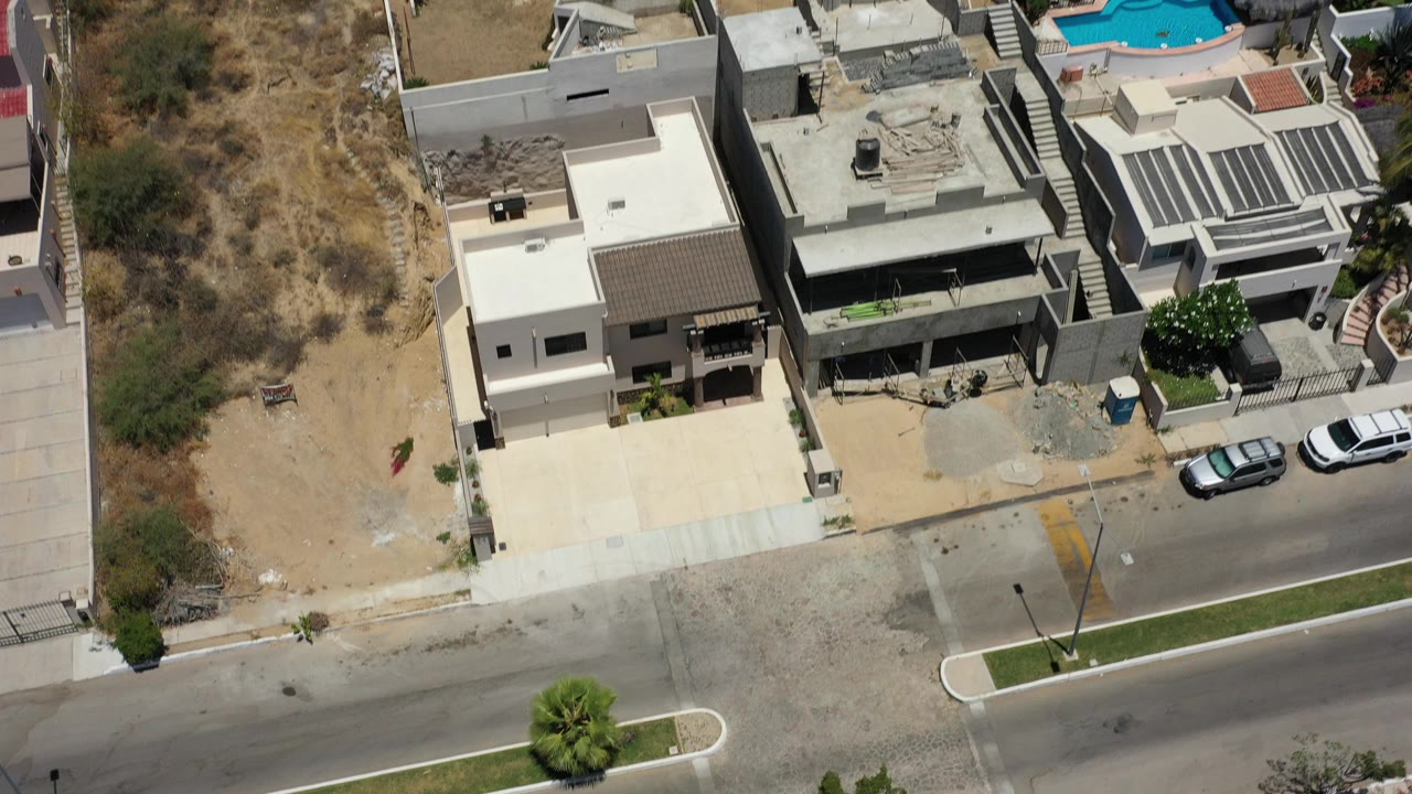 Opening overhead frame of modern homes and condos in San Jose del Cabo beside a paved road, dry vacant lot, parked cars, and no visible people.
