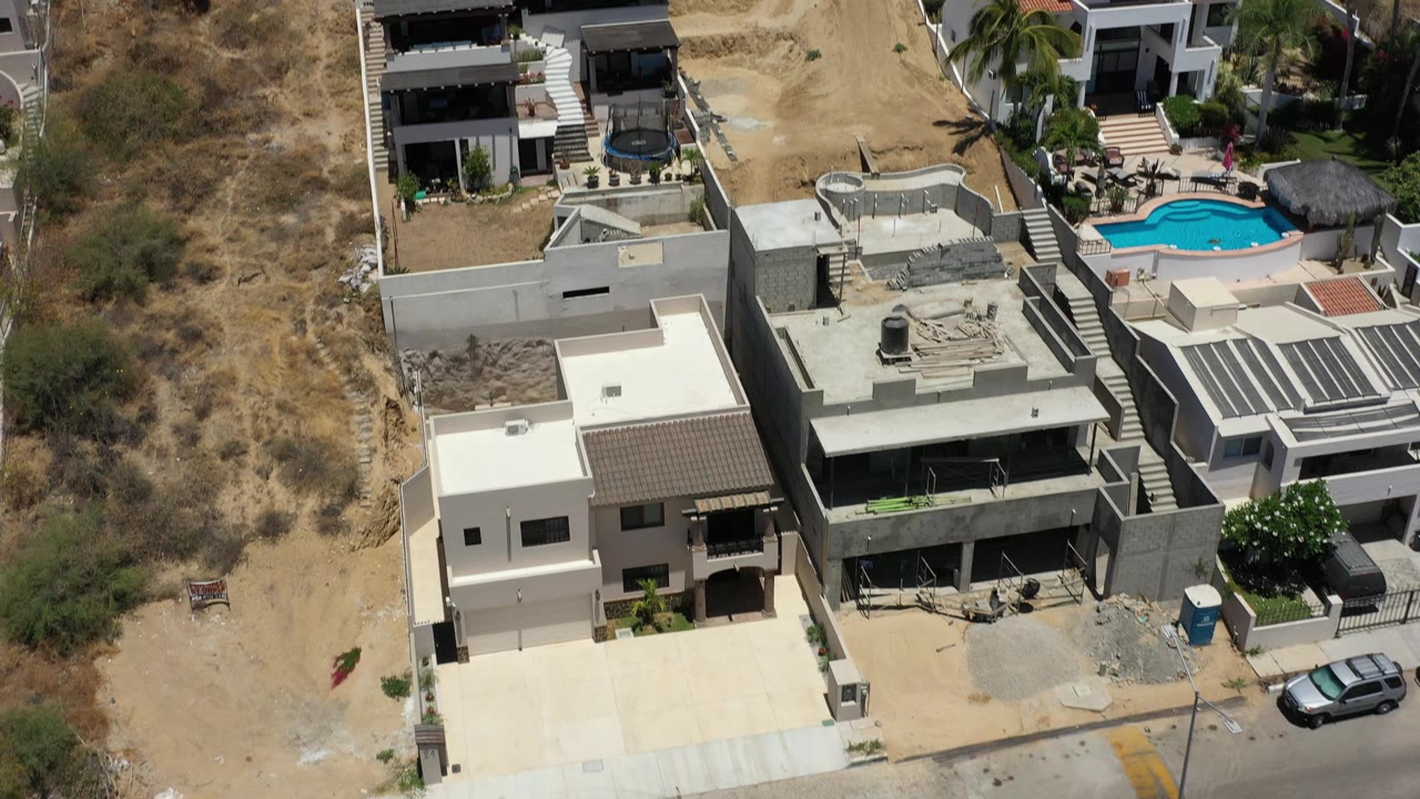 Aerial view in San Jose del Cabo, Mexico, centered on flat-roofed homes with a dry hillside to the left, a small pool beyond the houses, parked vehicles, and no visible people.