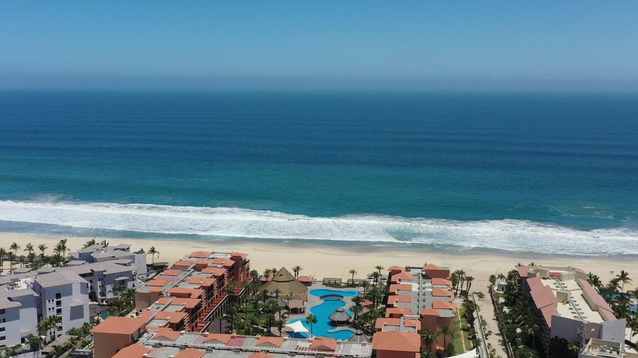Aerial frame over San Jose del Cabo, Mexico, with deep blue ocean beyond a white surf line and red-roofed coastal homes, palms, and bright pools in the foreground, no visible people.