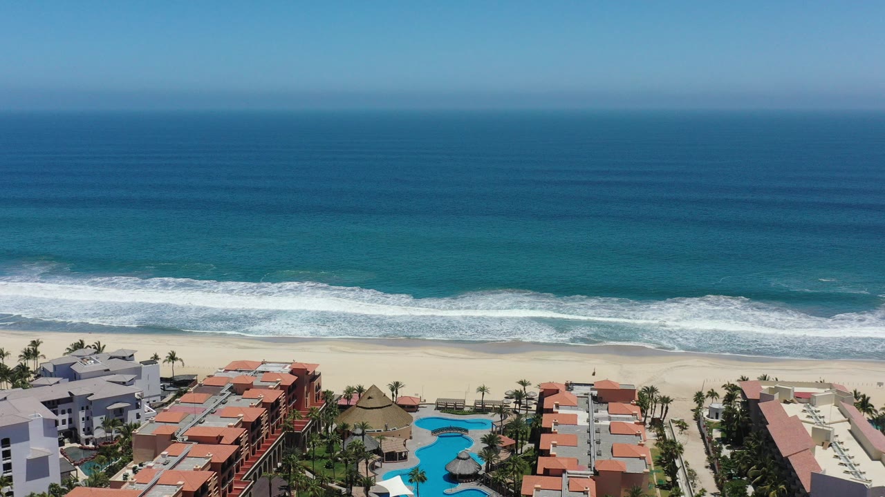 Aerial frame over San Jose del Cabo, Mexico showing deep blue ocean, a white surf line, pale sand, and red-roofed coastal buildings with turquoise pools below.