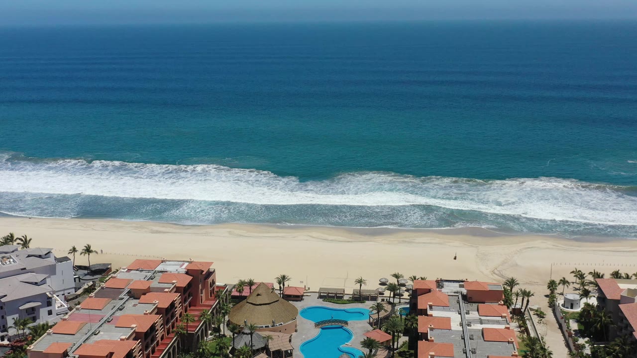 Aerial view in San Jose Del Cabo, Mexico with a broad turquoise ocean band, a foamy wave line near shore, pale sand, resort-style buildings, pools, palms, and no visible people.