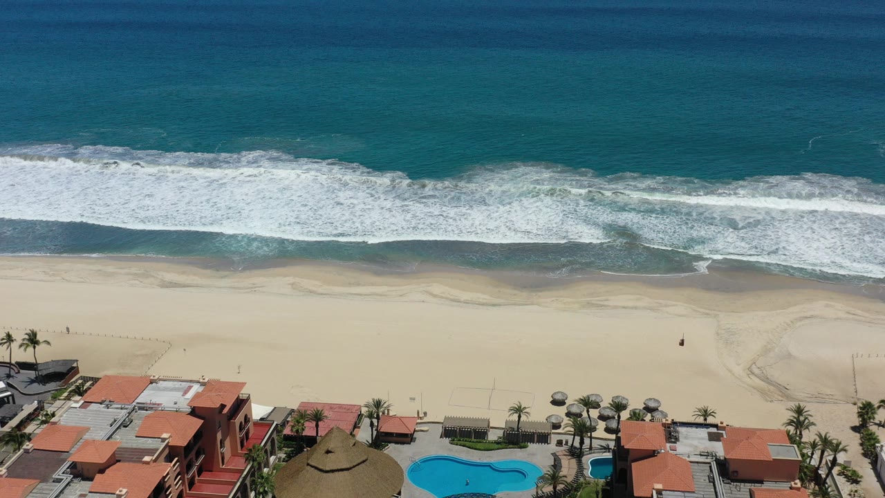 Aerial beach view with surf, sand, resort pools, and red-roofed buildings.
