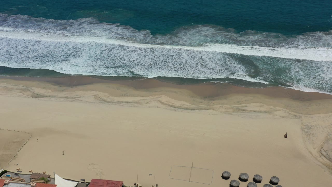 Top-down aerial view of white surf meeting tan sand in San Jose del Cabo, Mexico, with small beach structures along the lower edge and no visible people.