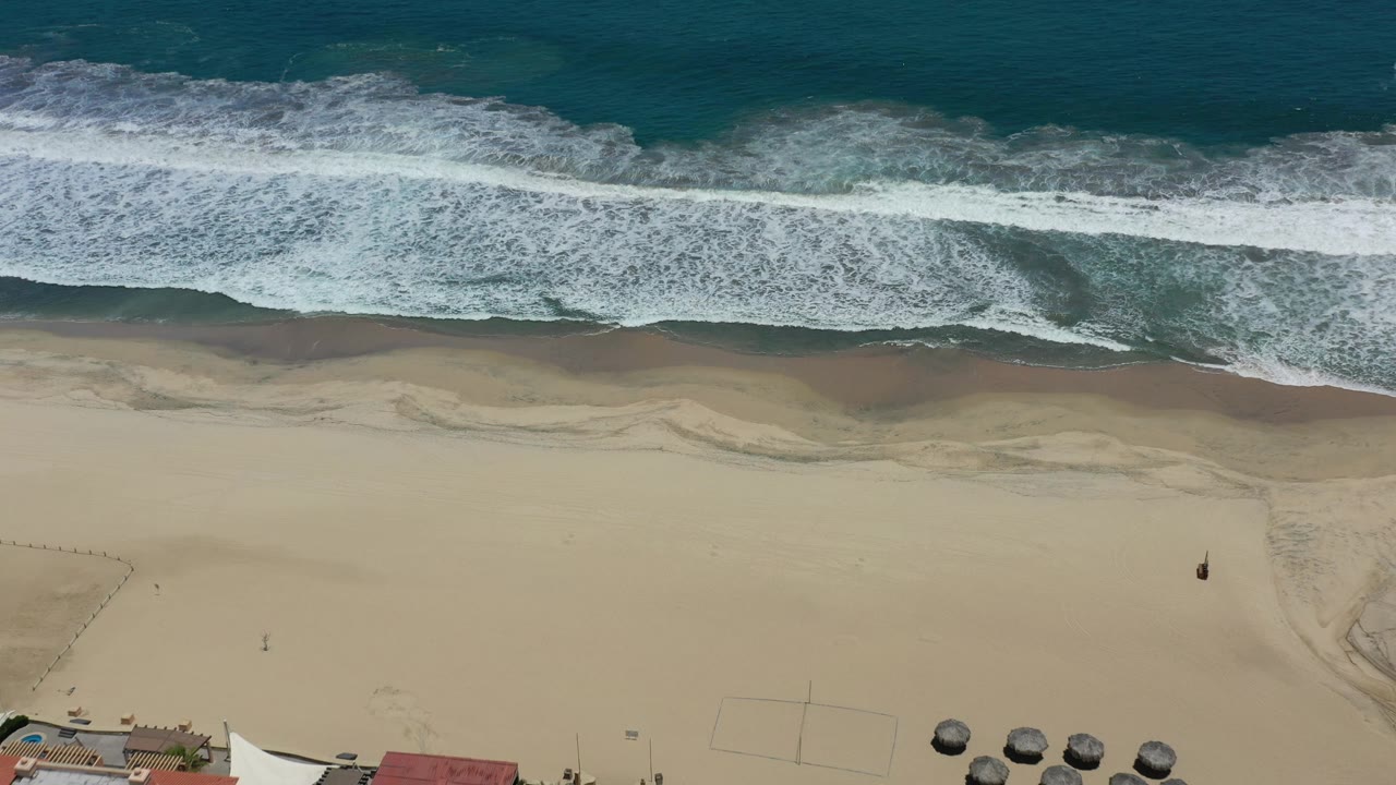 High overhead view of the San Jose del Cabo shoreline as foamy waves spread across darker wet sand, with open beach below and no visible people.