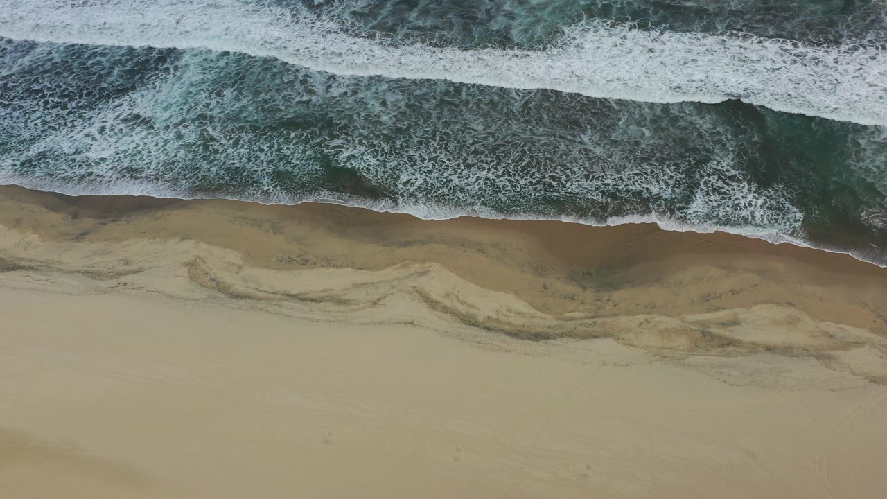 Overhead frame from San Jose Del Cabo, Mexico showing white surf at the top, dark green water, tan wet sand, and no visible people.