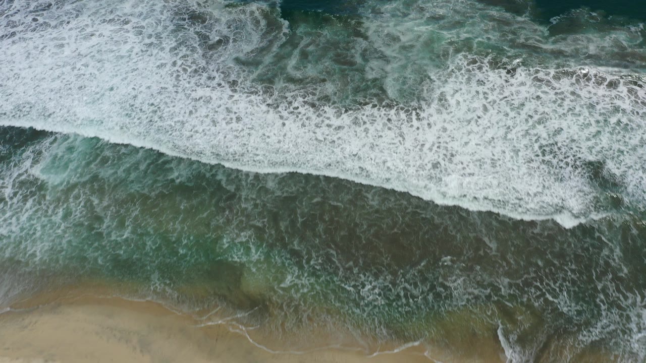 Overhead view of teal surf and white foam meeting a sandy shoreline in San Jose Del Cabo, Mexico, with no visible people.