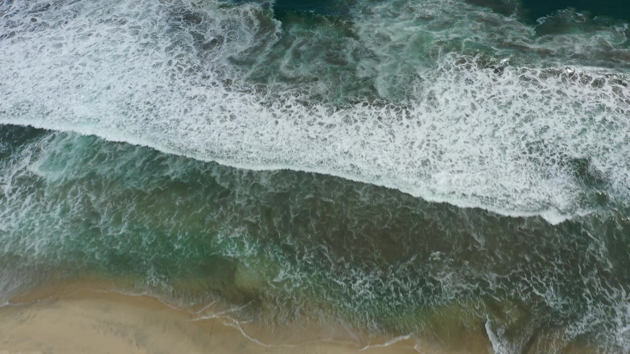 Aerial frame of white surf bands spreading across green-blue coastal water near sand in San Jose Del Cabo, Mexico, with no visible people.