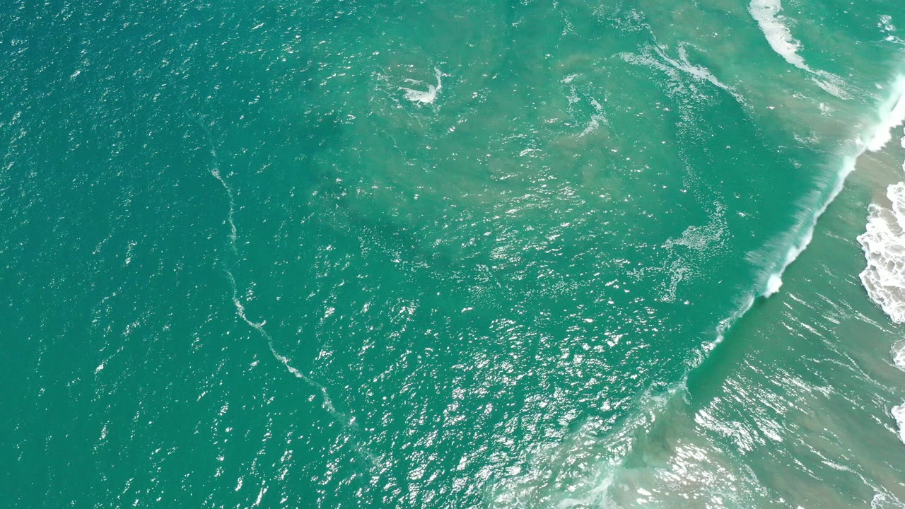 Blue-green ocean surface in San Jose del Cabo, Mexico, with bright sun sparkle and a diagonal white wave near the right side, no visible people.