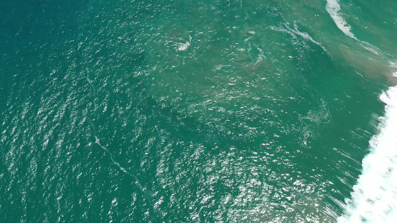 Overhead view of turquoise ocean in San Jose del Cabo, Mexico, with bright sun sparkle across ripples, a pale shallow band near the top, white surf at the right edge, and no visible people.