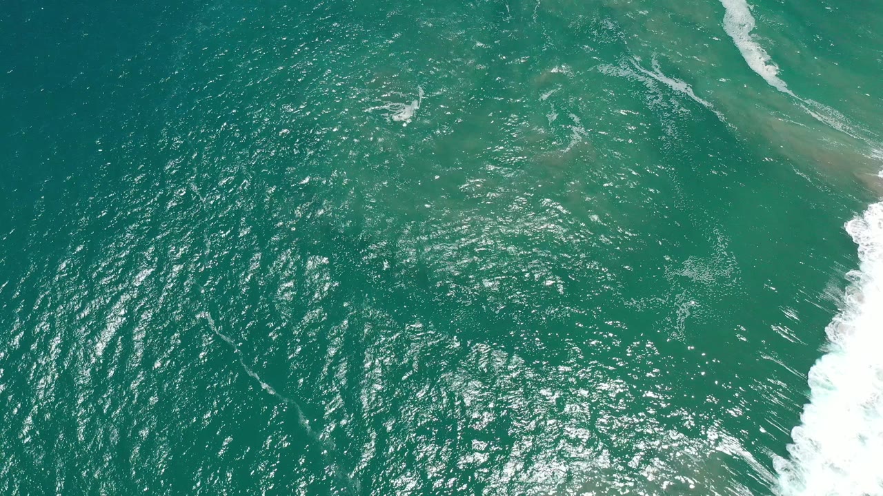 Top-down view over teal Pacific water in San Jose del Cabo, with dense sun reflections across the center, faint shallow color changes near the top, white waves at right, and no visible people.