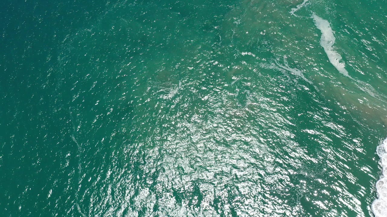 Overhead view of green ocean water in San Jose del Cabo, Mexico, with bright surface reflection and a thin pale wave band at the right edge, no visible people.