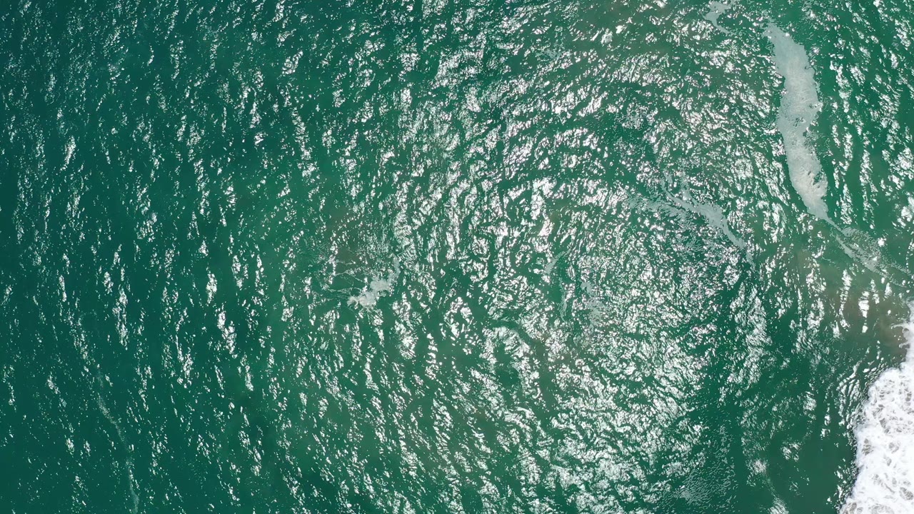 Aerial-style view of San Jose del Cabo ocean water with dense silver highlights across the ripples and a vertical strip of foam on the right, with no visible people.