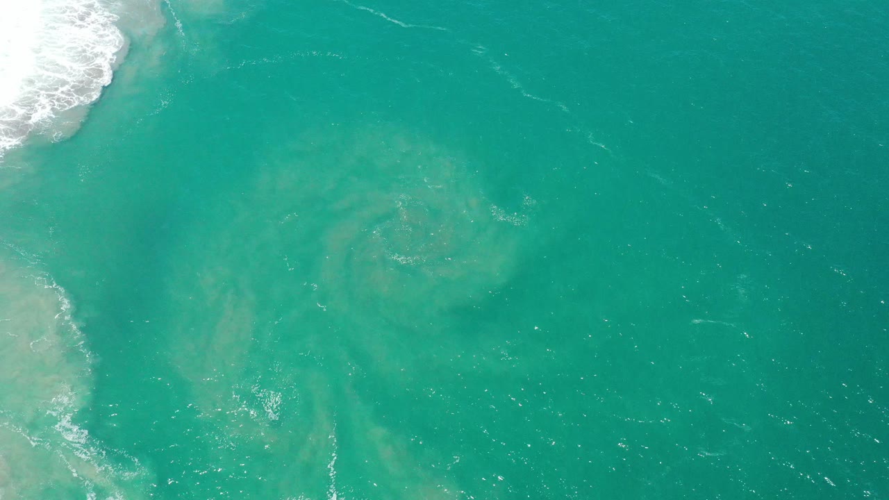 Overhead San Jose del Cabo ocean frame with a pale swirl spreading through turquoise water, white surf on the left edge, and no visible people.