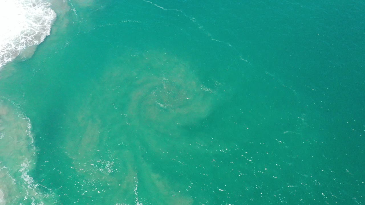 Overhead view of green-blue seawater in San Jose del Cabo with pale sandy swirls below the surface, white surf at the upper left, and no visible people.