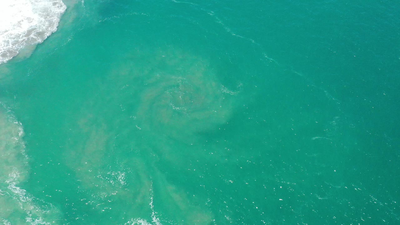 Top-down view of teal ocean water in San Jose Del Cabo, Mexico, with pale circular clouding below the surface and white surf foam at the upper left, no visible people.