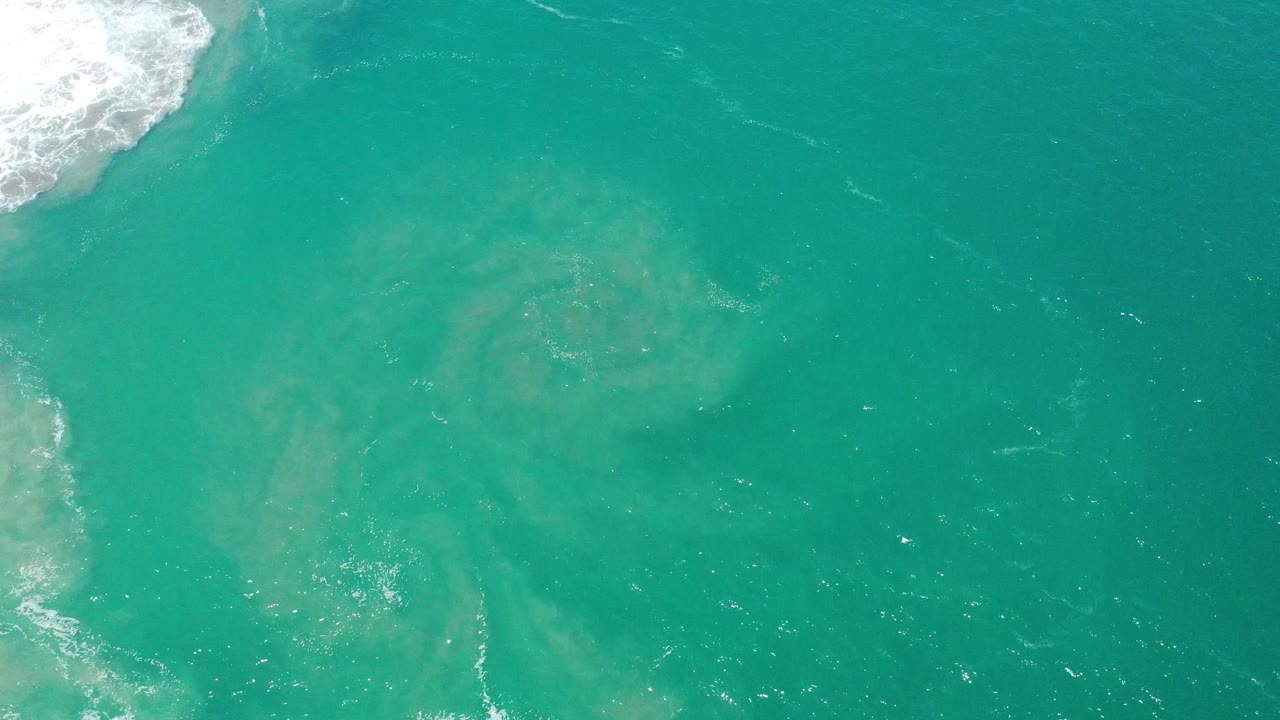Top-down teal ocean surface with a light circular patch and foam near the upper left.