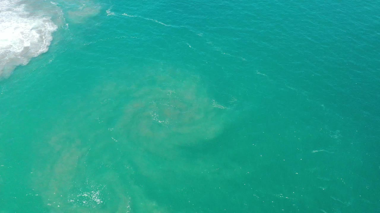 At 0:01, teal water near San Jose Del Cabo shows a larger milky tan swirl below the surface, small ripples, a bright reflection patch, and no visible people.