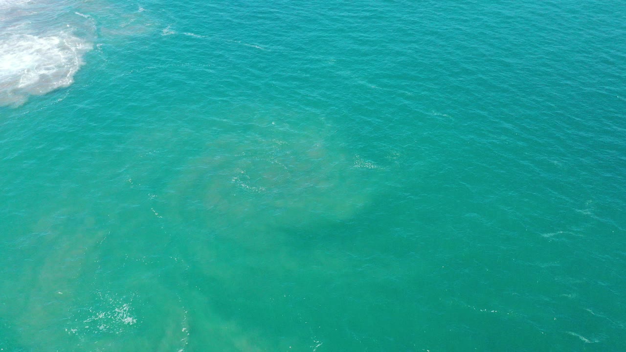 Turquoise shallow sea water in San Jose del Cabo shows faint sand suspended below the surface, soft ripples, and a patch of white surf foam at the upper left, with no visible people.