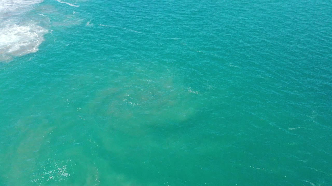 Rippling turquoise water with suspended sand patches below and a small surf edge at upper left.