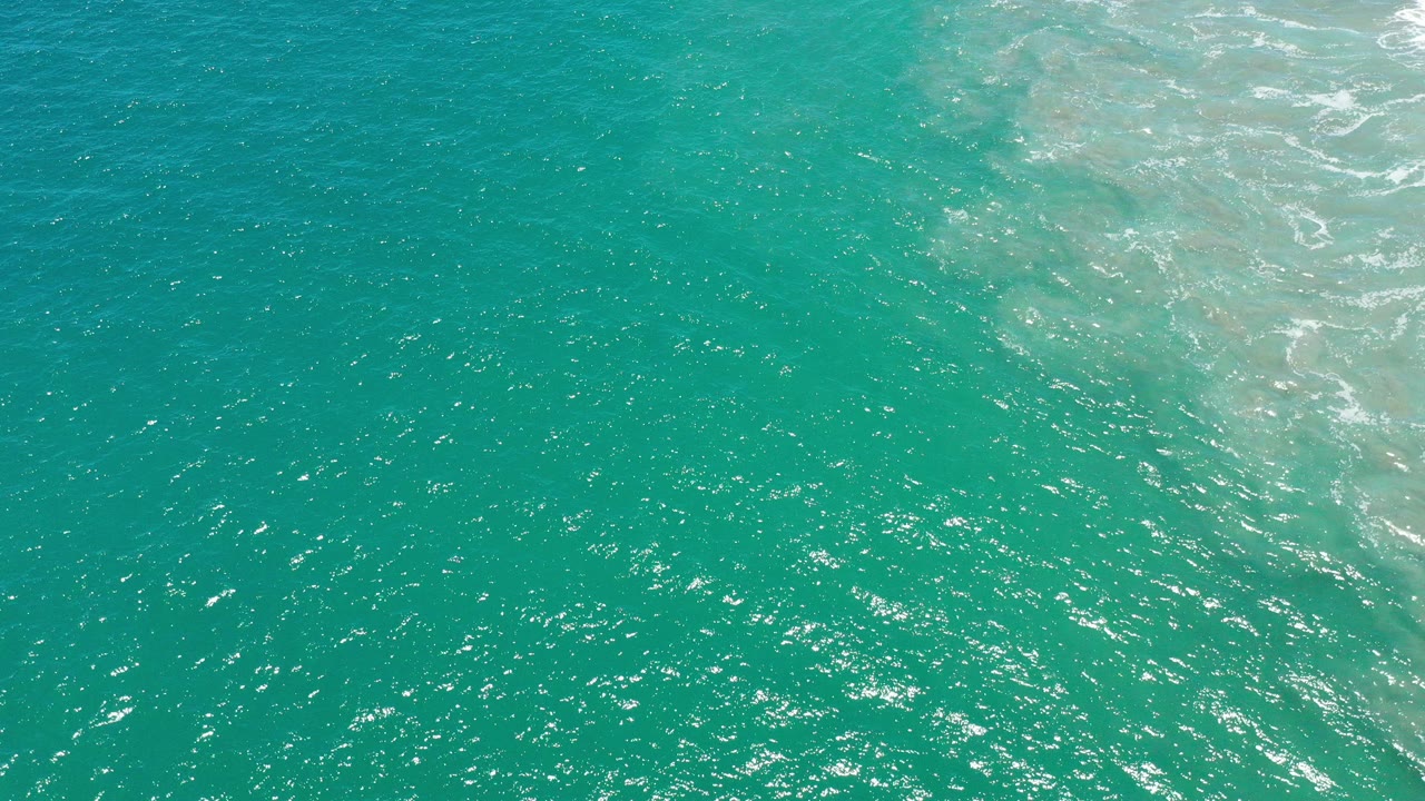 Top-down view of turquoise ocean water in San Jose del Cabo, Mexico, with scattered sun highlights and a pale foam band at the right edge, with no visible people.
