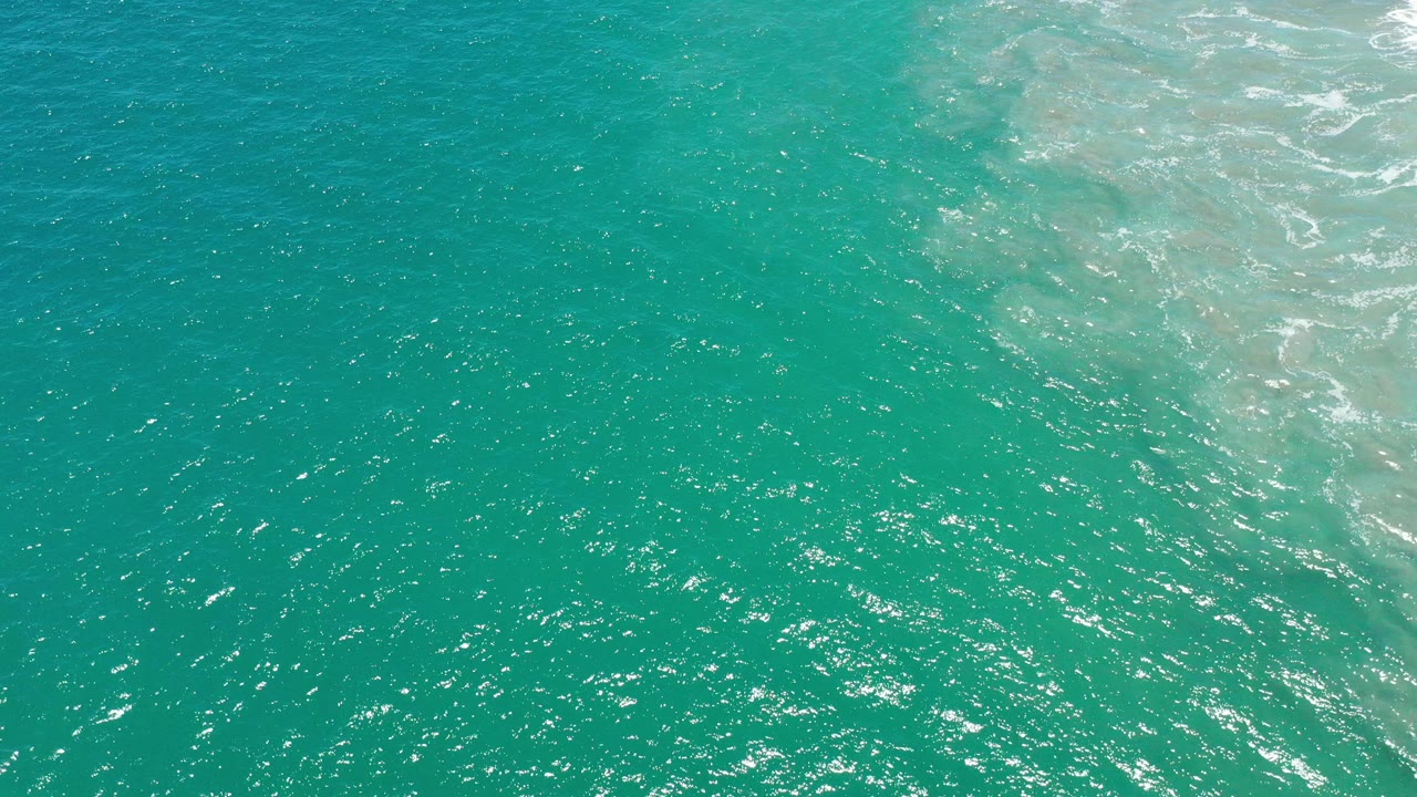 Aerial water detail over San Jose del Cabo, Mexico, showing aqua ripples, bright speckled reflections, and a lighter surf patch near the upper right, with no visible people.