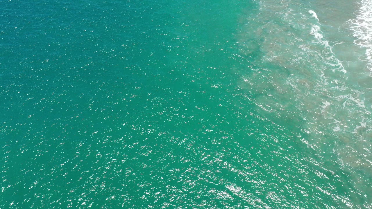 Overhead view of sea-green water in San Jose del Cabo with bright surface ripples, a pale shallow patch, white surf at the upper right, and no visible people.