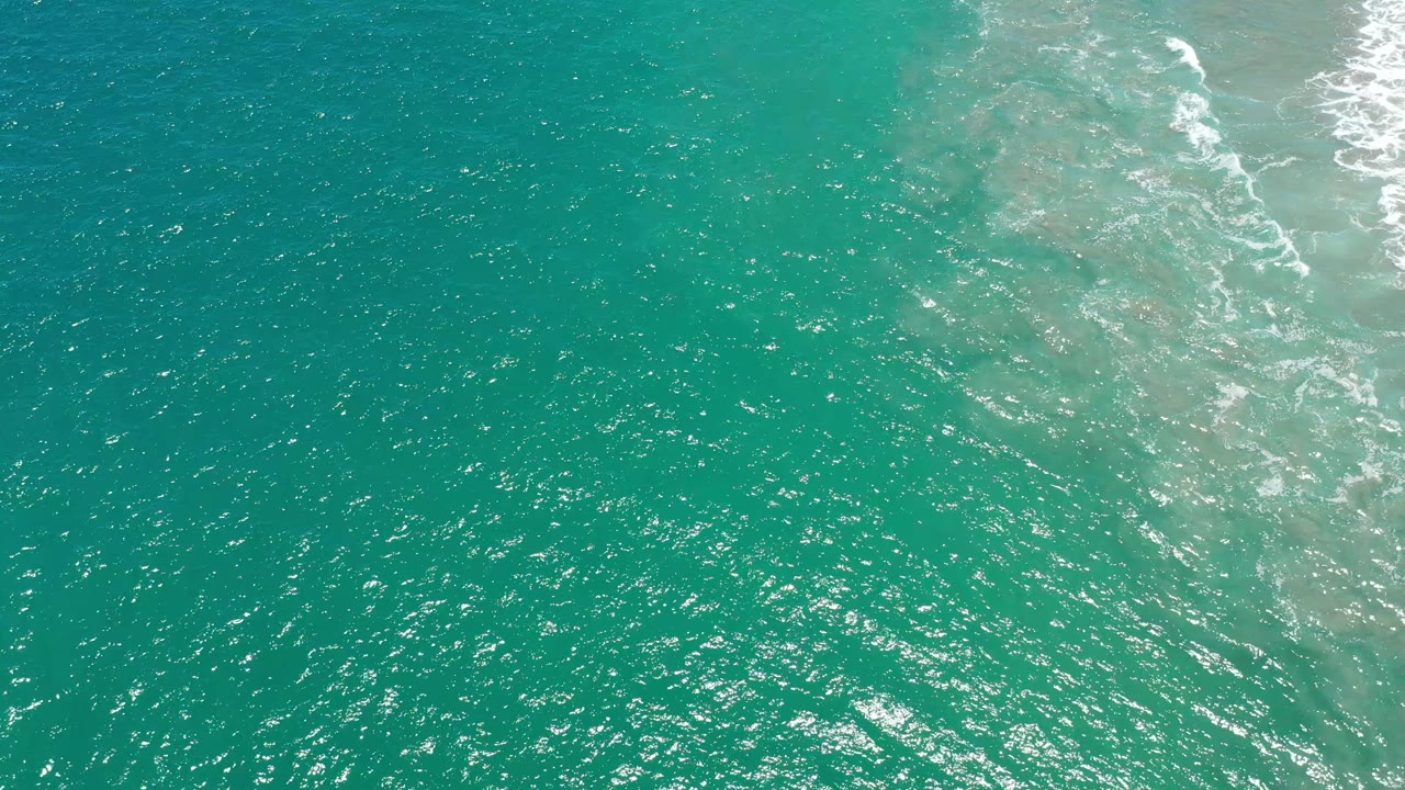 Aerial-looking frame of San Jose del Cabo ocean water with fine ripples across the center, lighter sandy tones below the surface, white foam near the top right, and no visible people.